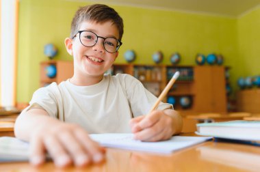 Gözlük takan, ders çalışan ve bir not defterine yazı yazan mutlu bir çocuk.