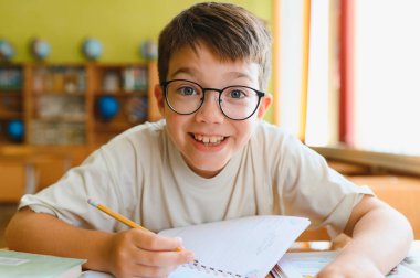Gözlüklü mutlu bir ilkokul öğrencisi not defterine yazıyor, sınıfta çalışıyor ve öğreniyor.