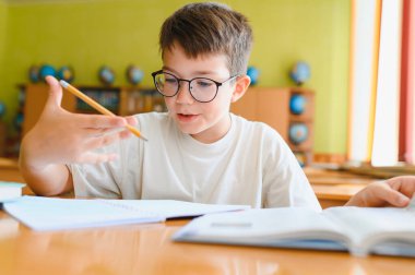 Gözlüklü genç bir çocuk, bir okul masasında öğrenmeye odaklanmış, ders kitabını okurken bir kalem tutuyor ve bilgiyi özümsüyor.