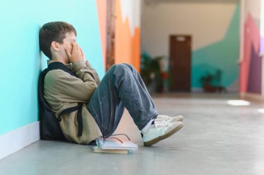 Üzgün okul çocuğu okul koridorunda yerde tek başına oturuyor ve sınıf arkadaşlarının zorbalığından acı çektikten sonra elleriyle yüzünü kapatıyordu.