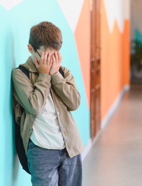 Okulda zorbalıktan muzdarip olan okullu çocuk elleriyle yüzünü örtüyor ve koridordaki duvarın yanında ağlıyor.