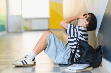 Üzgün okul çocuğu koridorda yerde oturuyor ve sınıf arkadaşlarının zorbalığı yüzünden ağlıyor.