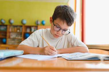 Gözlüklü genç bir öğrenci ödevlerine konsantre oluyor ve bir sınıfta deftere yazı yazıyor.