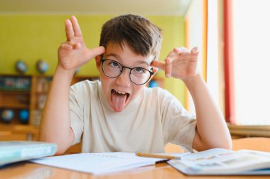 Genç bir çocuk gözlük takıyor komik bir yüz ifadesi takıyor ve okulda bir masada otururken dilini dışarı çıkarıyor.