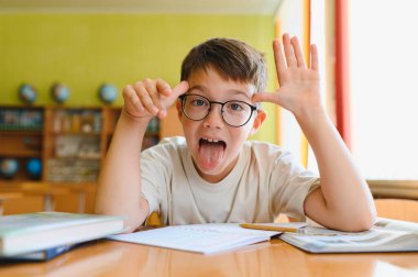 İlkokul öğrencisi, kitap ve okul malzemeleriyle çevrili bir masada çalışırken komik bir ifade takınıyor, neşe ve neşe saçıyor.