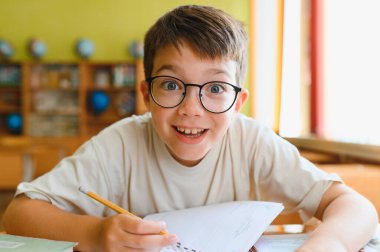 Gözlük takan mutlu ilkokul öğrencisi okul masasında deftere yazıyor, okula geri dönüyor.