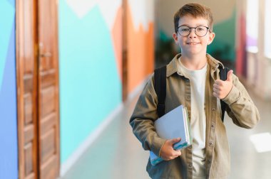 İlkokul öğrencisi renkli bir okul koridorunda yürürken elinde kitaplarla başparmağını kaldırıyor.