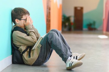 Okul koridorunda yerde oturup ağlayan, sınıf arkadaşlarının zorbalığından acı çektikten sonra yüzünü elleriyle örten üzgün öğrenci.
