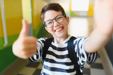 Sırt çantalı mutlu çocuk baş parmak işareti yapıyor, parlak bir koridorda okul zamanının tadını çıkarıyor.