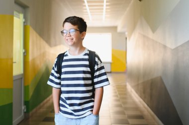 Mutlu çocuk gözlük takıyor ve sırt çantası taşıyor. Dersten sonra canlı bir okul koridorunda yürürken gülümsüyor.