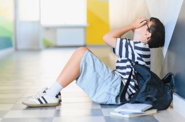 Öğrenci çocuk koridorda yerde oturuyor, sınıf arkadaşlarının zorbalığından acı çektikten sonra ağlayarak ve elleriyle yüzünü örtüyor.