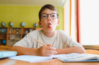 Gözlüklü genç, hayat dolu bir sınıfta ödevlerine konsantre olurken bir yandan da dilini dışarı çıkartıyor.