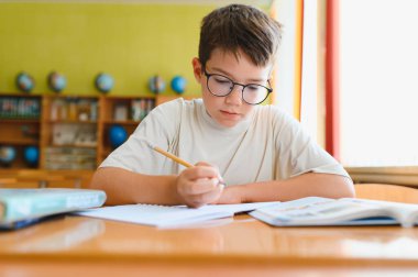 Gözlüklü genç okul ödevinde özenle çalışıyor, sınıftaki masada not defterine yazıyor.