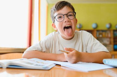 Masada oturup ders çalışırken dilini dışarı çıkaran oyuncu bir ilkokul öğrencisi.