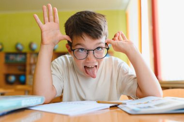 Liseli çocuk, neşeli bir sınıfta masasında otururken dilini çıkarıp komik bir ifade takınıyor.