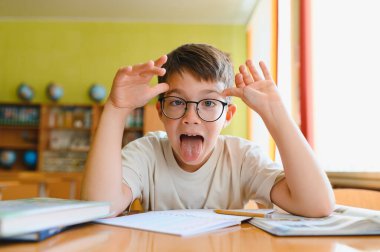 Liseli çocuk, sınıftaki masasında otururken dilini çıkarıp komik bir ifade takınıyor.