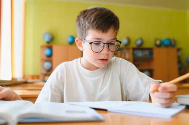 Gözlüklü genç öğrenci, bir sınıf masasında otururken öğrenmeye ve özenle bir not defterine yazmaya odaklanır.