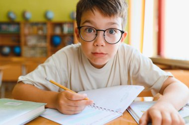 Gözlük takan zeki çocuk ders çalışıyor, not defterine kurşun kalemle okul masasında yazıyor.