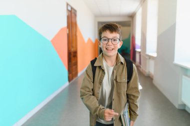 Renkli okul koridorunda kitap ve sırt çantası taşıyarak yürüyen mutlu bir ilkokul öğrencisi.