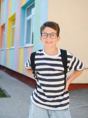 Mutlu çocuk renkli bir okulun yanında dururken göz kırpıyor ve gülümsüyor, okul zamanının heyecanıyla eğleniyor.