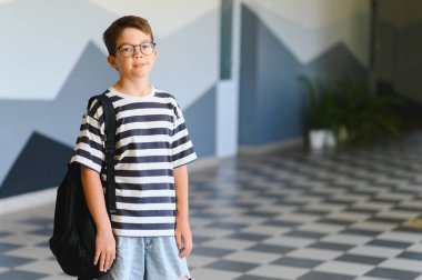 Gözlüklü ve sırt çantalı gülümseyen bir öğrencinin portresi. Okul koridorunda duruyor. Öğrenmeye ve eğitime hazır.