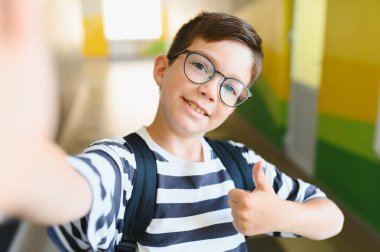 Gözlüklü mutlu öğrenci, selfie çekiyor ve okul koridorunda baş parmak işareti yapıyor. Öğrenme sevinci ve coşkusu yayıyor.