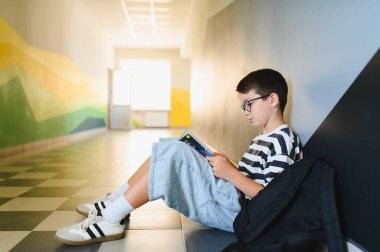 Gözlüklü bir çocuk okul koridorunda oturmuş, sırt çantasıyla ders kitabı okuyor, öğrenme üzerine odaklanmış.