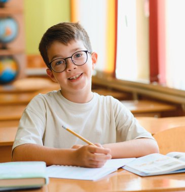 Mutlu bir ilkokul öğrencisi kurşun kalemle bir not defterine yazıp parlak bir sınıfın masasında oturarak öğrenmeye odaklanmış.