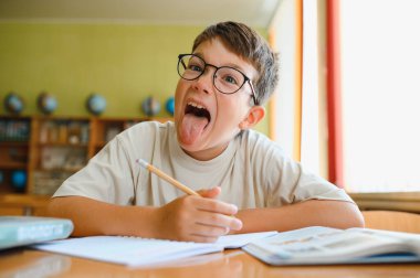 Gözlüklü genç, ödevlerine dalmışken ve okul masasında ders çalışırken dilini şakayla uzatıyor.