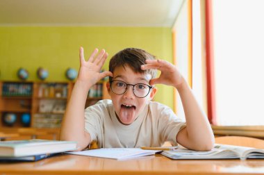 Bir sınıftaki masada otururken yüzünü buruşturan ve dilini dışarı çıkaran oyuncu bir öğrenci.