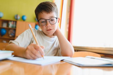 Gözlüklü genç okul ödevinde özenle çalışıyor, sınıftaki masada not defterine yazıyor.
