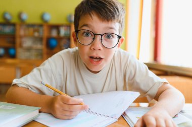 Gözlüklü ilkokul öğrencisi sınıfta defterine yazarken şaşırmış bir ifade takınıyor.