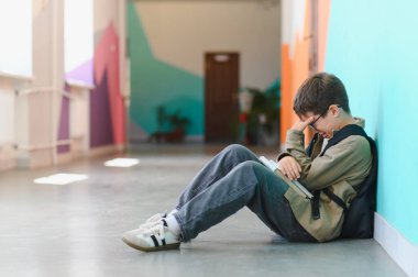 Sınıf arkadaşlarının zorbalığına uğradıktan sonra okulda yerde ağlayan üzgün okul çocuğu elinde bir kitap tutuyor ve sırt çantası var.