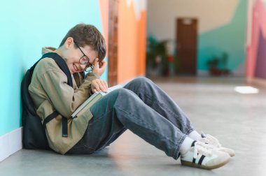 Okul koridorunda yerde oturan, yüzünü kitapla örten ve sınıf arkadaşlarının zorbalığı yüzünden ağlayan üzgün öğrenci.