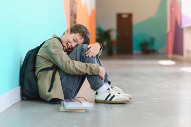 Okulda zorbalığa uğradıktan sonra sırt çantası ve kitaplarla yerde oturup ağlayan üzgün okul çocuğu.