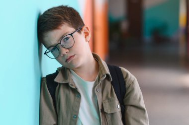 Sırt çantalı bir öğrenci duvara yaslanmış, sınıf arkadaşlarının zorbalığına katlanıyor, üzüntü ve yalnızlığı yansıtıyor.