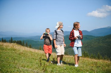 Bir grup çocuk dağ manzarasında yürüyüş yapmaktan zevk alıyor, doğayı keşfediyor ve dürbün ve kamera kullanıyor.