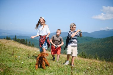 Sırt çantalı bir grup çocuk çimenli dağ yamacında yürüyorlar tasmalı küçük bir köpekle, yaz tatilinin tadını çıkarıyorlar.