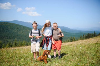 Sırt çantalı ve dürbünlü üç çocuk güneşli bir yaz gününde köpekleriyle dağ çayırında yürüyüş yapıyorlar.