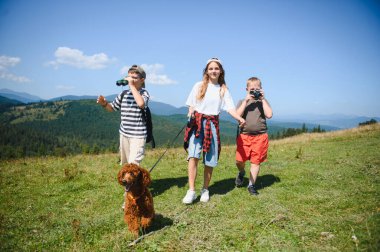 Bir grup çocuk köpekleriyle dağ yürüyüşünden zevk alıyor, dürbün ve kamerayla doğayı keşfediyorlar.