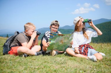 Üç genç yürüyüşçü, güneşli bir günde yolculuklarını sürdürmek için bir harita ve dürbünle çimenli bir dağın tepesinde oturuyorlar.