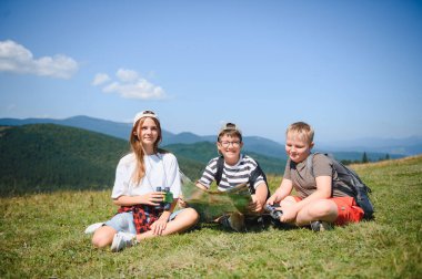 Üç genç yürüyüşçü dağlarda güneşli bir günün tadını çıkarıyor, haritaya danışıyor ve dürbün kullanıyorlar.