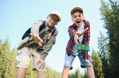 İki hevesli genç kaşif sırt çantaları ve dürbünleriyle yemyeşil bir ormanda yürüyüş yaparken izleyiciye doğru uzanıyorlar.