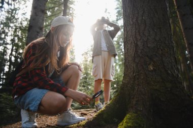 Doğayı keşfeden meraklı çocuklar, güneşli bir ormanda yürüyüş yaparken büyüteçle ağaç gövdesinde yosun gözlemliyorlar.