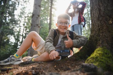 Meraklı çocuk, aile gezisi sırasında kız kardeşiyle birlikte dürbün kullanarak ormanı keşfederken baş parmağını kaldıran bir büyüteç kullanıyor.