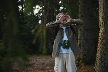 Genç kaşif, yoğun bir ormanda kaybolurken yardım için bağırıyor, dürbün tutuyor ve bir sırt çantası takıyor, bir çıkış yolu arıyor.