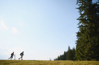 Canlı bir dağ çayırında koşan iki genç yürüyüşçü, yemyeşil ağaçlarla çevrili ve güneşli bir günün sıcaklığının tadını çıkarıyorlar.