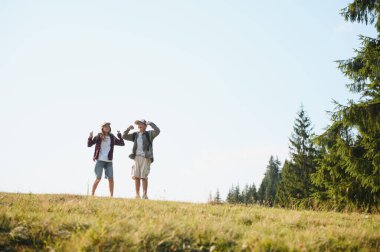 Bereketli orman ve parlak mavi gökyüzü ile çevrili bir dağ çayırının tepesinde başarılarını kutlayan iki neşeli izci.