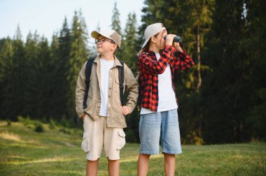 İki genç turist bir ormanı keşfediyor, biri dürbünle bakarken diğeri çevreyi gözetliyor.