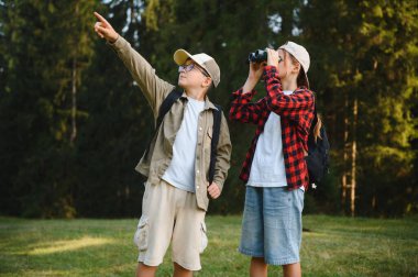 Doğayı keşfeden iki çocuk. Biri ilginç bir şeyi işaret ederken diğeri dürbün kullanıyor. Ormanda yürüyüş yapmaktan keyif alıyorlar.